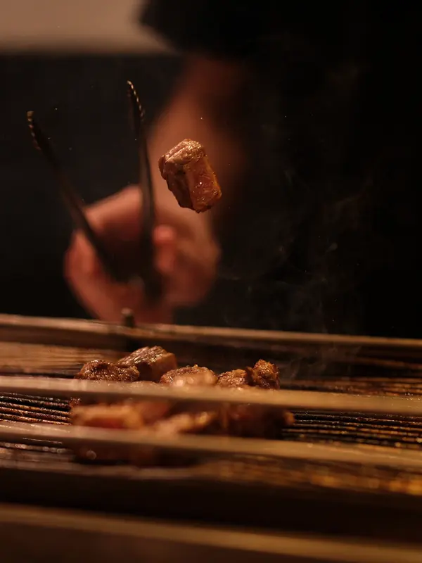 Mão do assador virando um pedaço de carne na grelha com um pegador, em luz baixa.