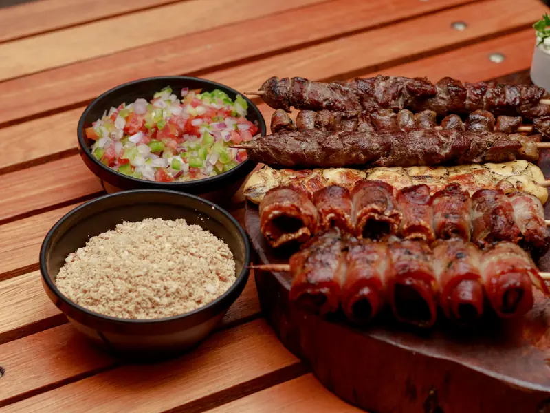 Espetos de carne sobre tábua de madeira, com tigelas de vinagrete e farofa.